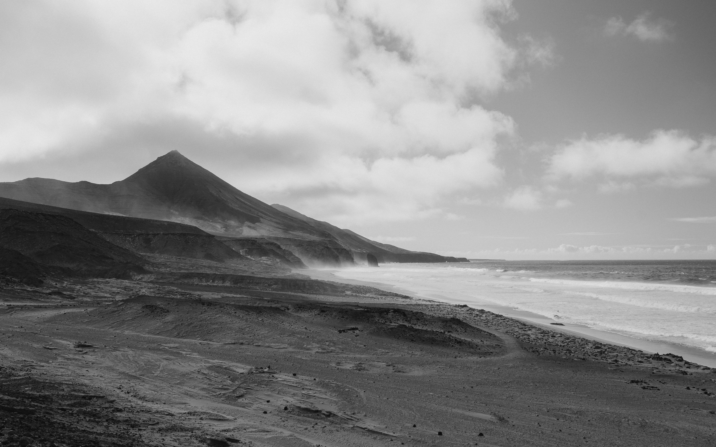 Black and white photo of a rugged coastline with a mountain in the background and waves crashing against the shore.
