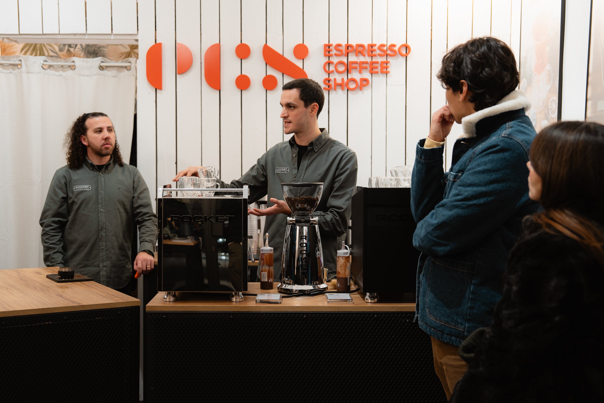 Barista explaining coffee to customers at a coffee shop counter with a sign that reads 'Espresso Coffee Shop' in the background.