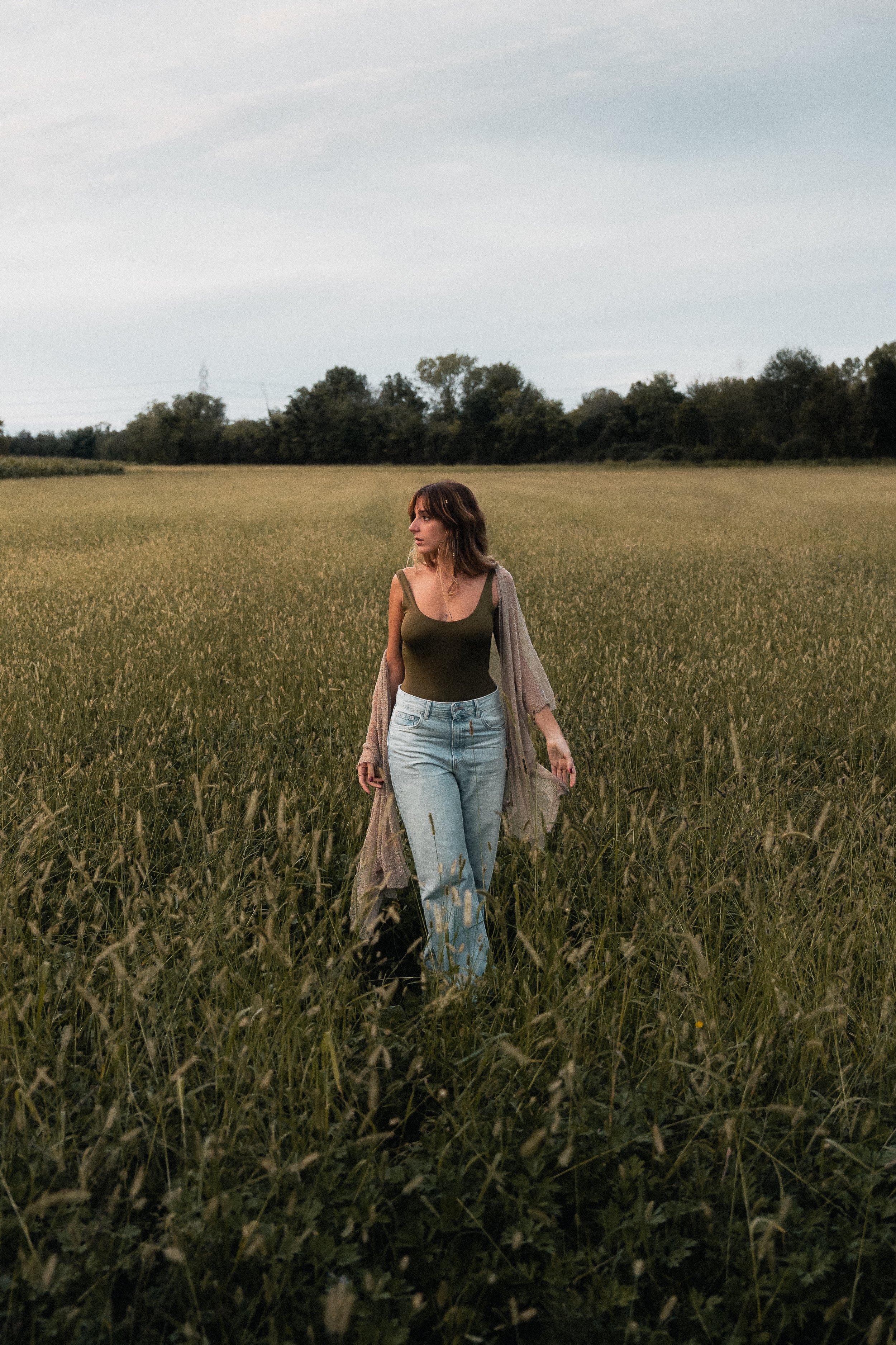 A woman walking through a grassy field during the daytime, wearing a green tank top, light blue jeans, and a long cardigan.