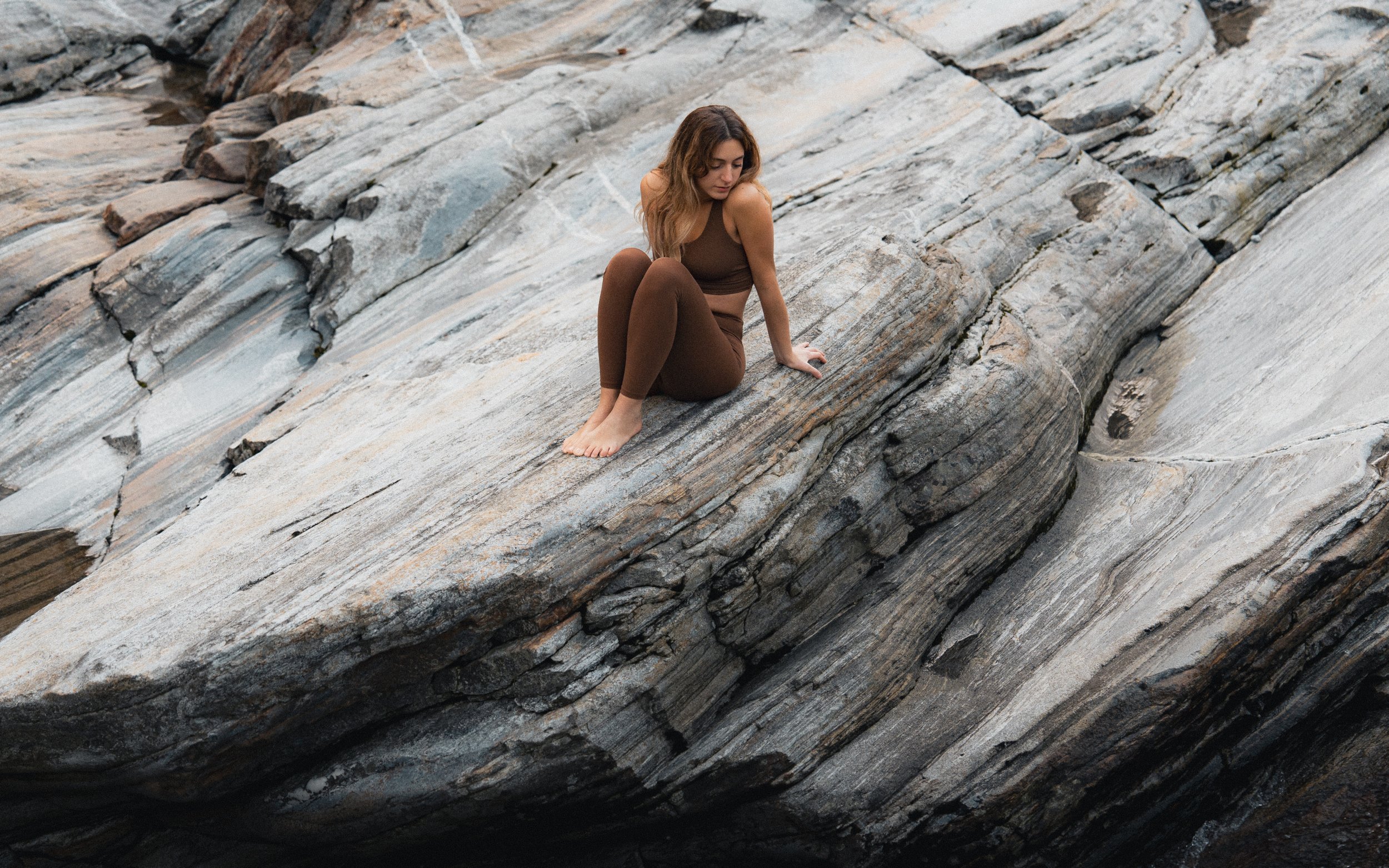 A woman sitting barefoot on a large, layered rock formation, wearing brown leggings and a sleeveless top, looking down thoughtfully.