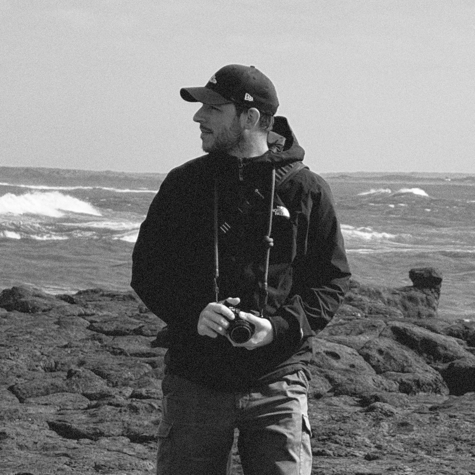 A man kneeling on rocks near the ocean, holding a camera, wearing a dark jacket, cap, and a backpack, with waves in the background.