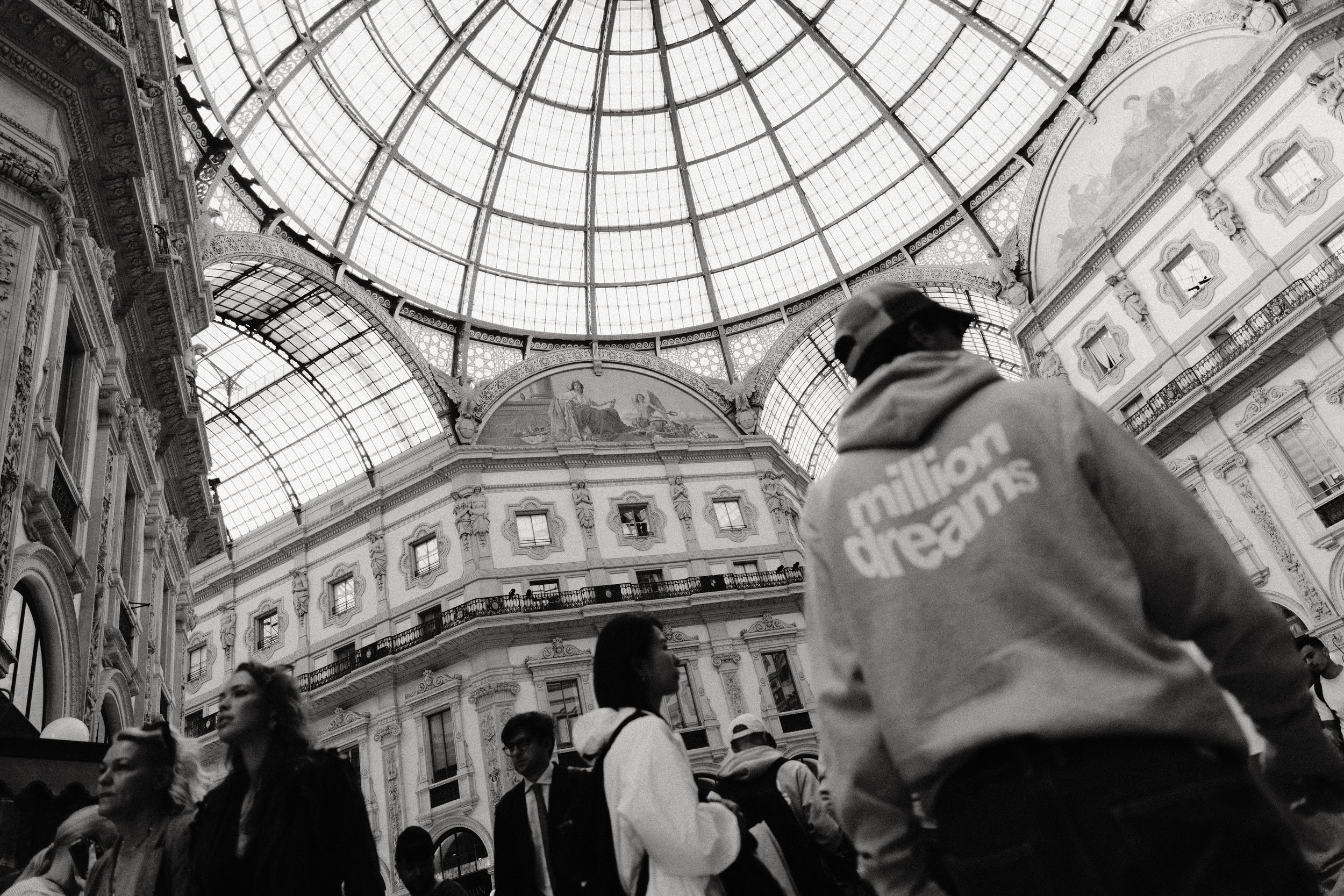 Multiple people walking inside a grand architectural building with a large glass dome ceiling and ornate decor.